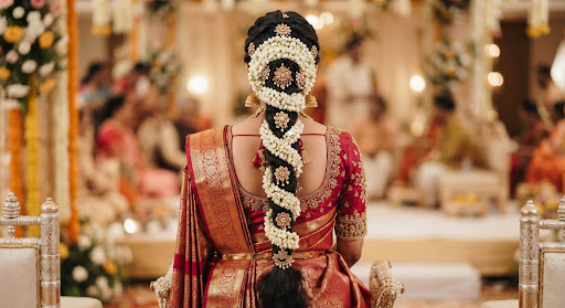 Rear angle of intricate South Asian bridal braided hair adorned with fresh white jasmine blooms gajra and golden hair accessories paired with royal crimson banarasi silk saree pleating and pallu draping.
